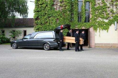 Ein Bestatterteam trägt einen Holzsarg aus einem Leichenschauhaus zu einem auf dem Parkplatz stehenden Bestattungswagen.