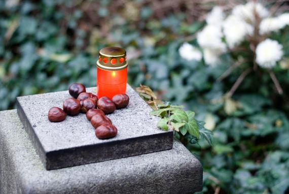 Rote Grabkerze steht auf einem Stein, umgeben von Kastanien und grünen Blättern. Hintergrund mit verschwommenen Blüten.