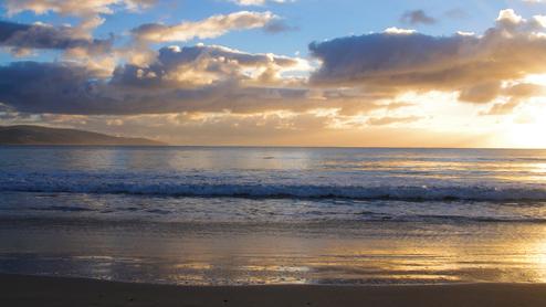 Sonnenuntergang über dem Meer, sanfte Wellen brechen am ruhigen Strand, Wolken reflektieren das Licht am Himmel.