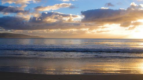 Sonnenuntergang über dem Meer, sanfte Wellen brechen am ruhigen Strand, Wolken reflektieren das Licht am Himmel.
