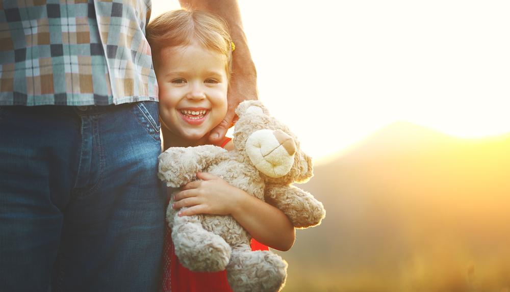 Lächelndes Kind mit einem Plüschteddy steht neben einem Erwachsenen, vor einer sonnigen, verschwommenen Landschaft.